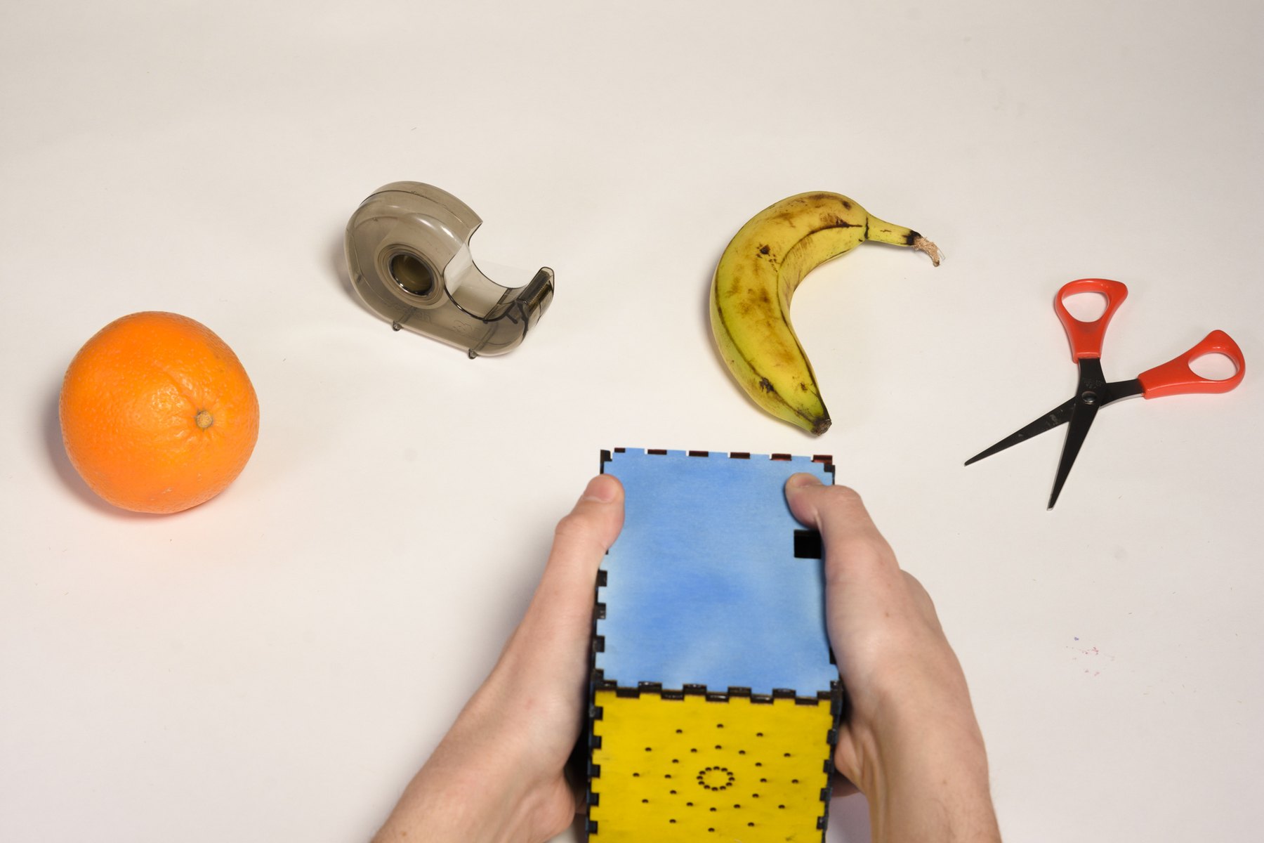Talki held in two hands, pointed at objects on a table: an orange, a tape dispenser, a banana, scissors